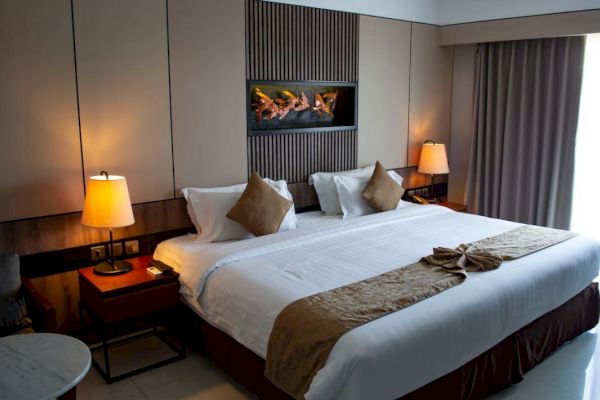 A well-lit, neatly arranged hotel room with a large bed, decorative pillows, two bedside tables with lamps, and a wall-mounted artwork above the bed.