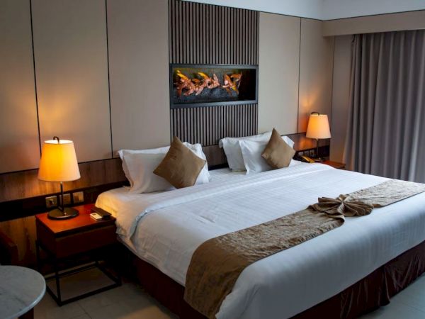 A well-lit, neatly arranged hotel room with a large bed, decorative pillows, two bedside tables with lamps, and a wall-mounted artwork above the bed.