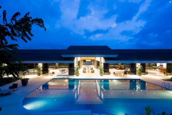 A modern, illuminated building with large open spaces and a swimming pool in front, set against a dusk sky.