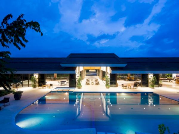 A modern, illuminated building with large open spaces and a swimming pool in front, set against a dusk sky.