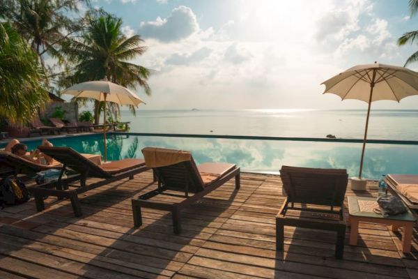 A serene poolside scene with lounge chairs, umbrellas, palm trees, and an ocean view under a sunny sky.