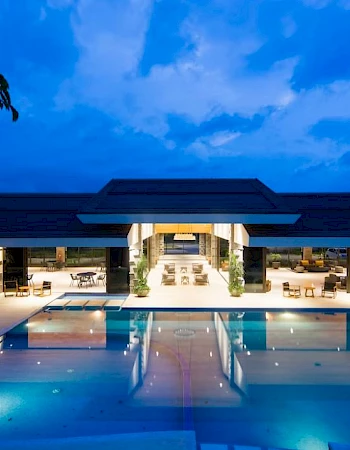 A luxurious open-air building with a large, illuminated swimming pool in the foreground, set against a twilight sky backdrop.
