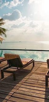 A serene poolside scene with lounge chairs and umbrellas on a wooden deck overlooking a calm body of water, surrounded by palm trees.