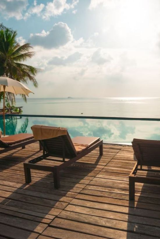 A serene poolside scene with lounge chairs and umbrellas overlooking calm waters, flanked by palm trees and a partly cloudy sky at sunrise or sunset.