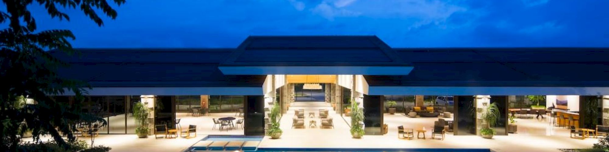 This image shows a modern building illuminated at night with a large, inviting swimming pool in the foreground, surrounded by lounge chairs.