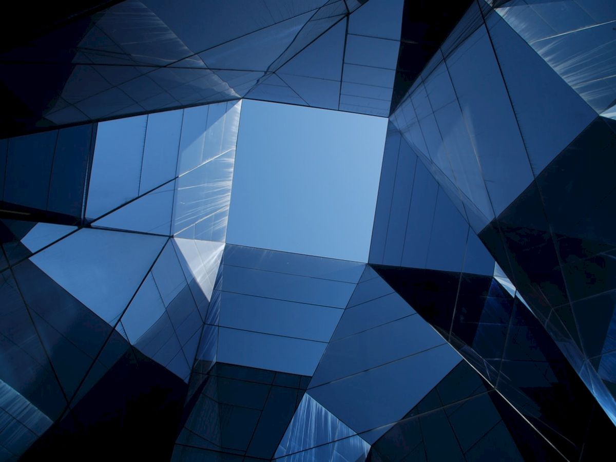 The image shows a geometric arrangement of reflective glass surfaces, with a clear blue sky visible through a polygonal opening in the center.