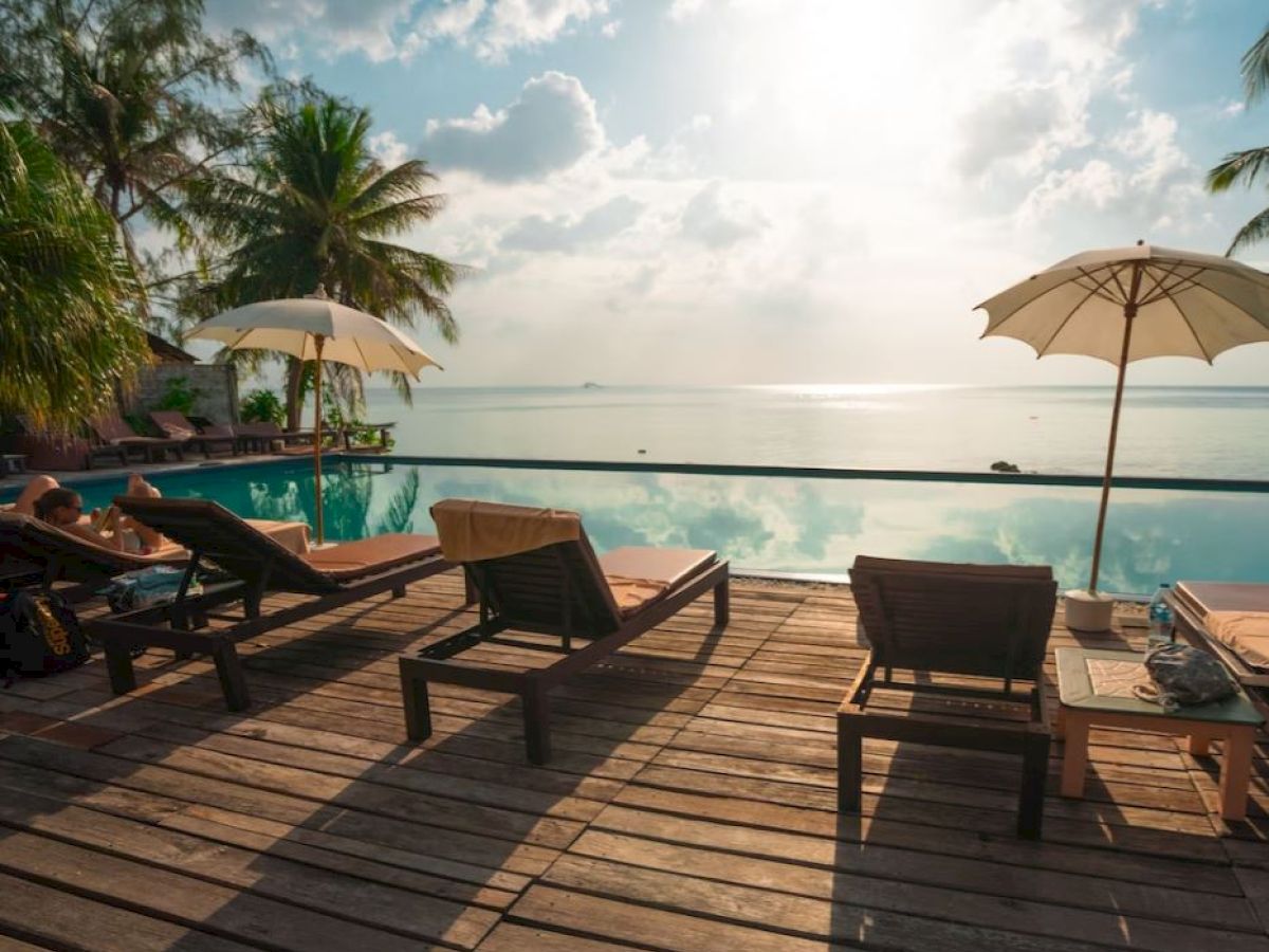 A peaceful poolside scene with lounge chairs, umbrellas, and palm trees overlooking a calm body of water under a bright, cloudy sky.