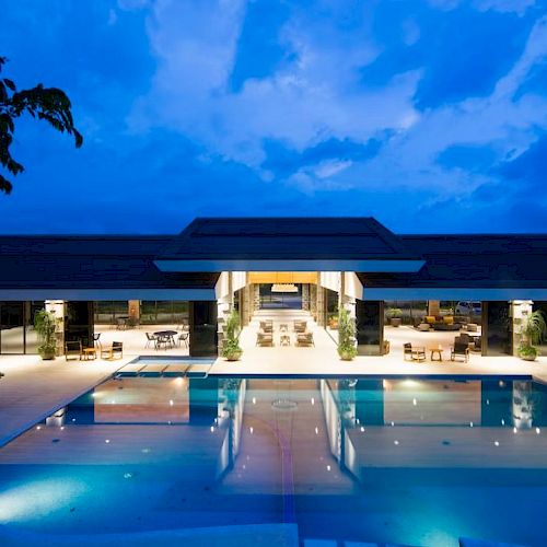 The image showcases a luxurious resort or villa with a large, illuminated swimming pool and spacious, modern architecture under a dusky blue sky.