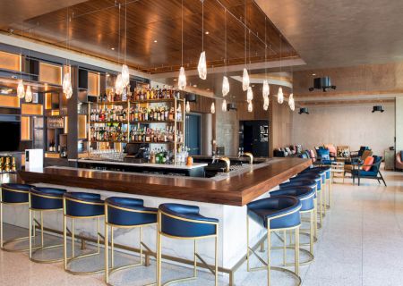 A modern bar with blue stools, a well-stocked back shelf, hanging lights, and a seating area with couches in the background.
