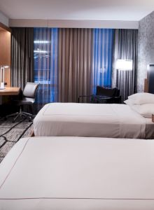 A hotel room features two neatly made beds, a TV, a work desk with a chair, modern lighting, and large windows with blinds.