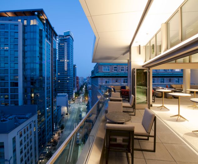 The image shows a modern cityscape with illuminated tall buildings and an expansive balcony, featuring tables and chairs, during twilight.