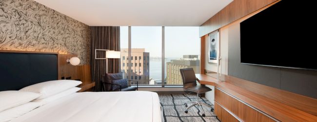 This image shows a modern hotel room with a large bed, wall-mounted TV, desk, chair, and a window with a city view.
