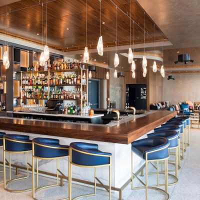 This image shows a modern bar with blue cushioned stools, pendant lights, and a well-stocked bar counter, located in a stylish lounge area.