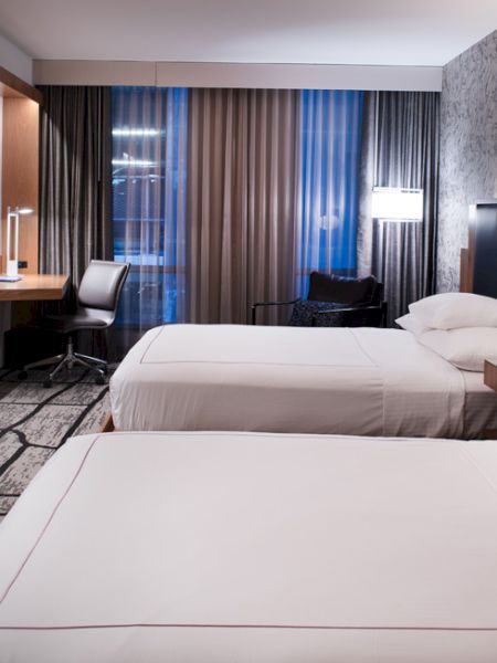 The image shows a modern hotel room with two neatly made beds, a wall-mounted TV, a desk with a chair, and stylish lighting fixtures.