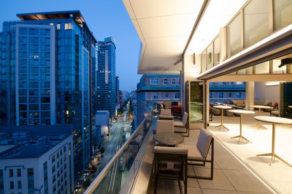 A modern balcony overlooks a city with tall buildings at dusk. The balcony has tables, chairs, and glass railings, with the city lights beginning to glow.