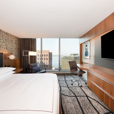 A modern hotel room with a king-size bed, large TV, desk with chair, decorative wall, and floor-to-ceiling windows showing a city view ends the sentence.