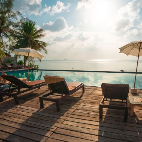 The image shows a serene beach or poolside setting with lounge chairs, umbrellas, and palm trees, overlooking calm waters and a beautiful sunny sky.