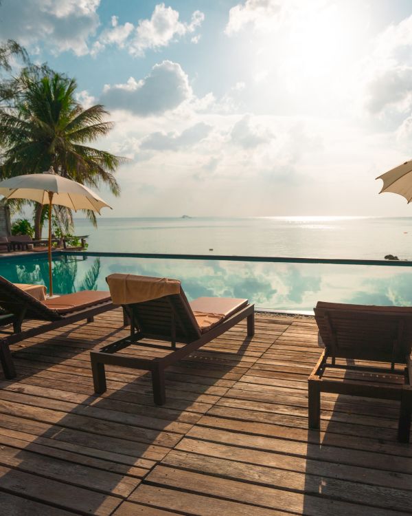 The image shows a serene beach or poolside setting with lounge chairs, umbrellas, and palm trees, overlooking calm waters and a beautiful sunny sky.