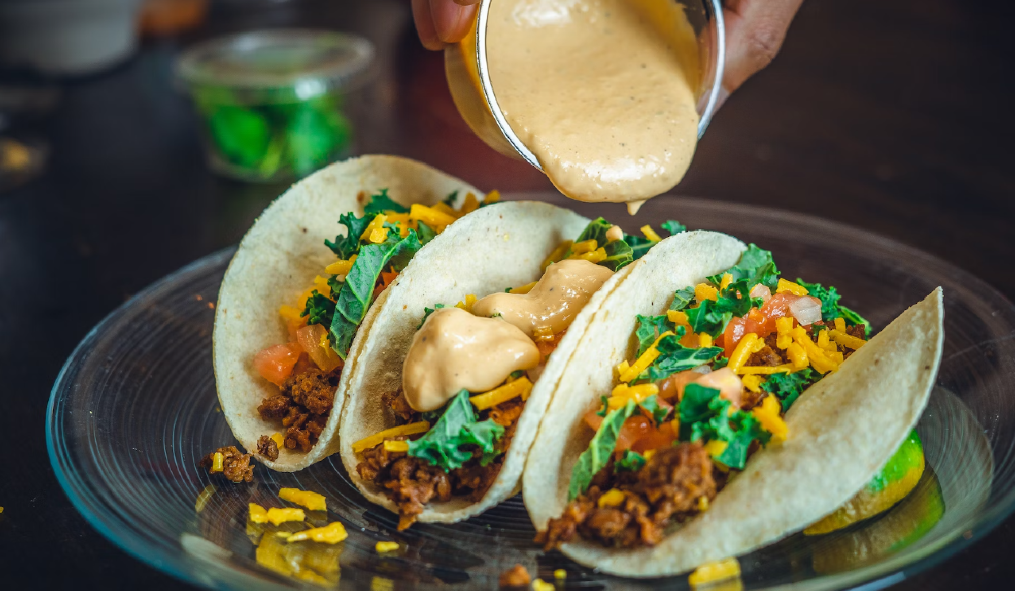 The image shows three tacos on a clear plate, being topped with a creamy sauce. The tacos contain lettuce, cheese, and diced tomatoes.