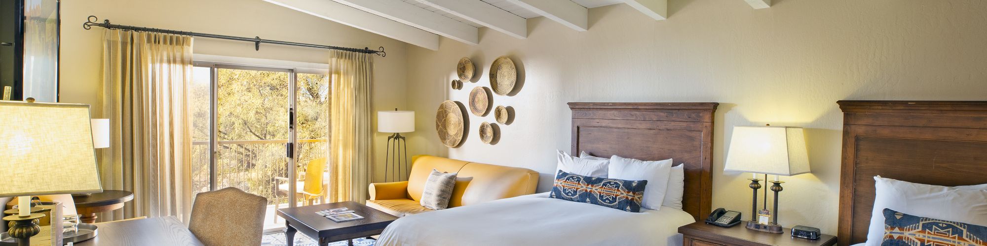 A spacious hotel room features two beds, a desk, a couch, a coffee table, and a sliding glass door leading to a balcony with outdoor scenery.