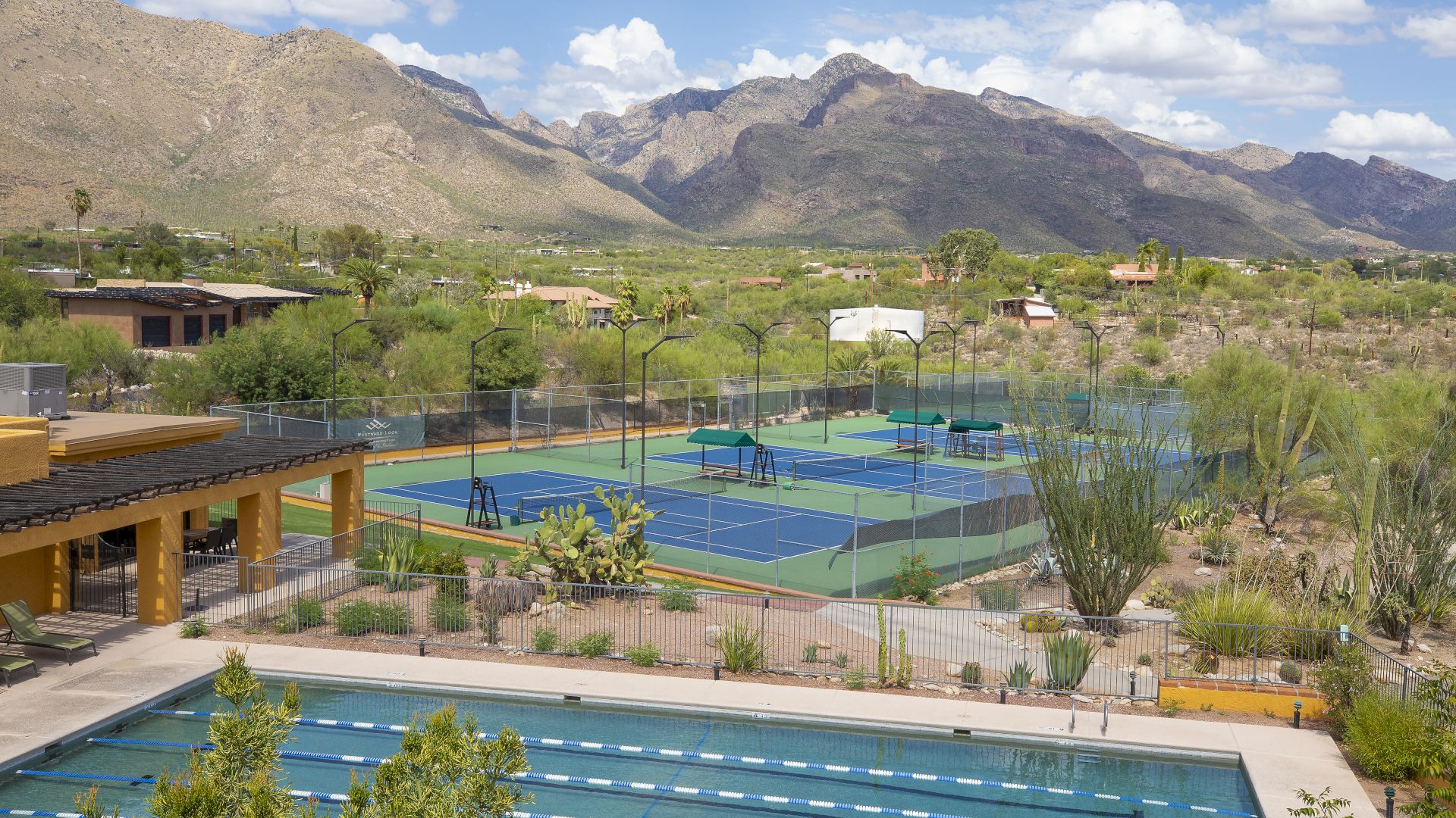 The image shows an outdoor recreational area with a swimming pool, multiple tennis courts, and mountainous scenery in the background, under a partly cloudy sky.