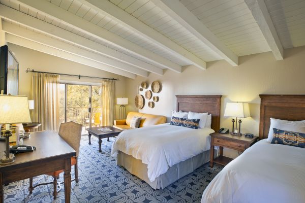 A cozy hotel room with two beds, a sofa, a desk, and a balcony view. Decor includes wall art, lamps, and patterned carpet under a beamed ceiling.
