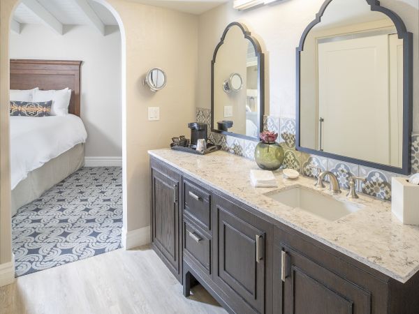 A modern bathroom with a double-sink vanity and large mirrors, connected to a bedroom through an arched doorway, featuring subtle decor and bright lighting.
