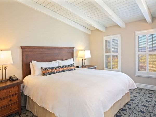 A cozy bedroom with a wooden bedframe, white bedding, bedside tables with lamps, a patterned rug, and two windows with white blinds.