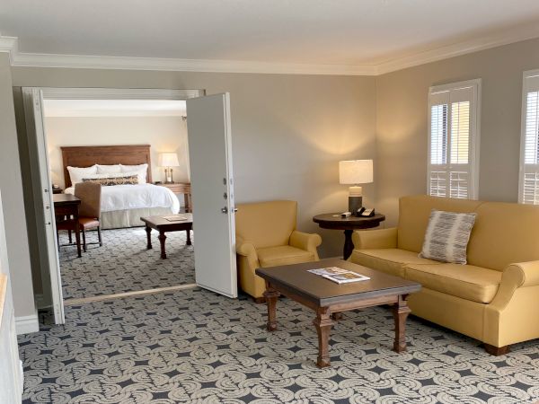 A cozy hotel room features a sitting area with a yellow couch, table, and lamp, and a bedroom with a large bed and two side tables visible through an open door.