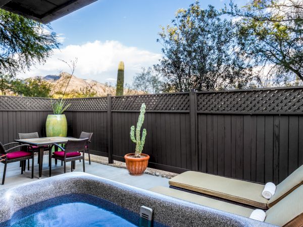 A cozy outdoor patio with a hot tub, dining table, chairs, potted plants, and mountain views, bordered by a wooden fence and lush greenery.