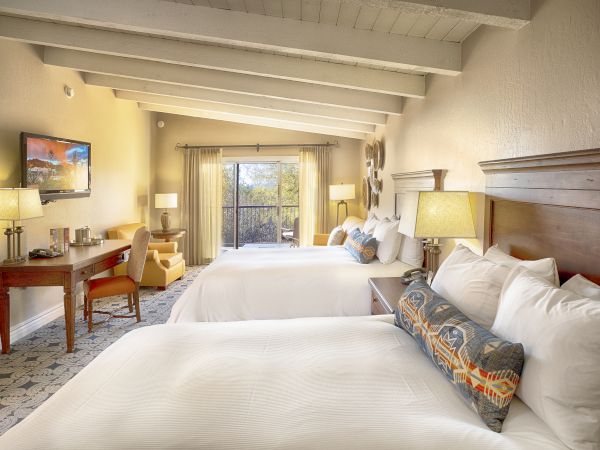 A cozy hotel room with two large beds, a desk with a chair, a TV, and a balcony view with natural light.
