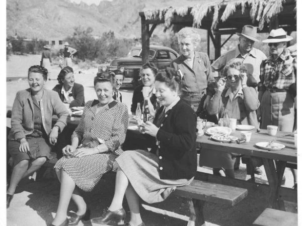 A group of people is gathered outdoors, sitting and standing around a picnic table with food and drinks, smiling and socializing in a sunny setting.