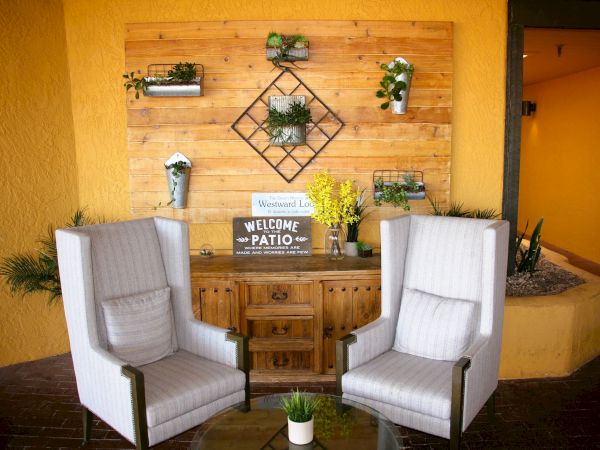 Cozy patio with two cushioned chairs, a wooden table, and a decorative wall with plants and a "Welcome to our Patio" sign.