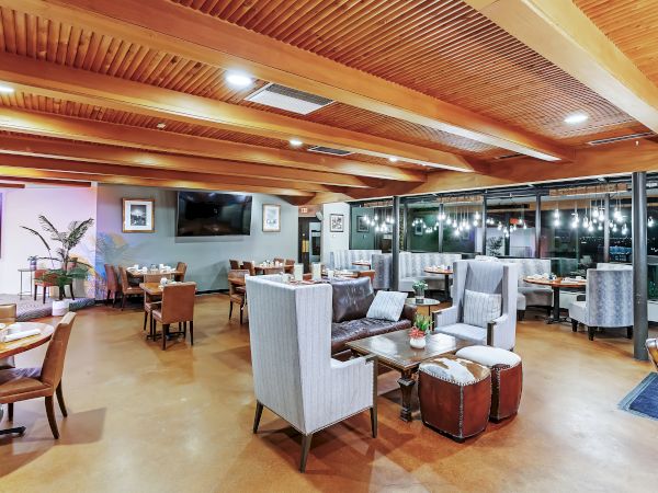A modern, well-lit restaurant interior with wooden ceilings and a mix of table and lounge seating, decorated with plants and wall art.