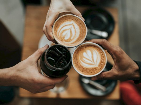 Three people holding cups of coffee, two with latte art and one with a dark beverage, are toasting over a table with plates and utensils.
