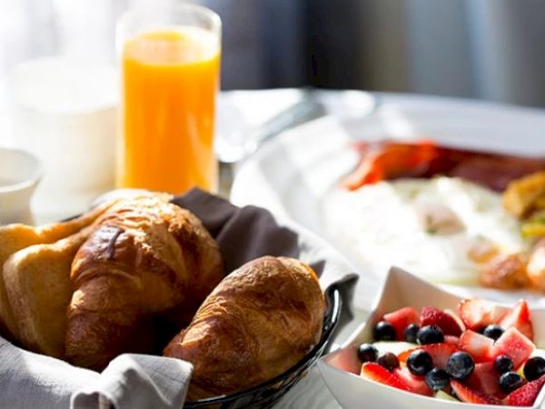 A breakfast spread with croissants, orange juice, a bowl of fruit, and a plate of eggs, bacon, and what looks like a toasted piece of bread.