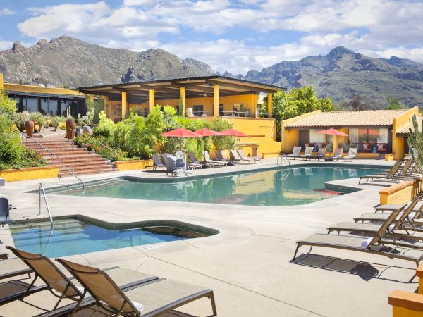 An outdoor pool area with lounge chairs, a hot tub, colorful umbrellas, and a view of mountains in the background is visible.