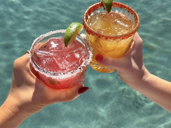 Two colorful cocktails with lime wedges raised in a sunny poolside toast, shimmering water below.
