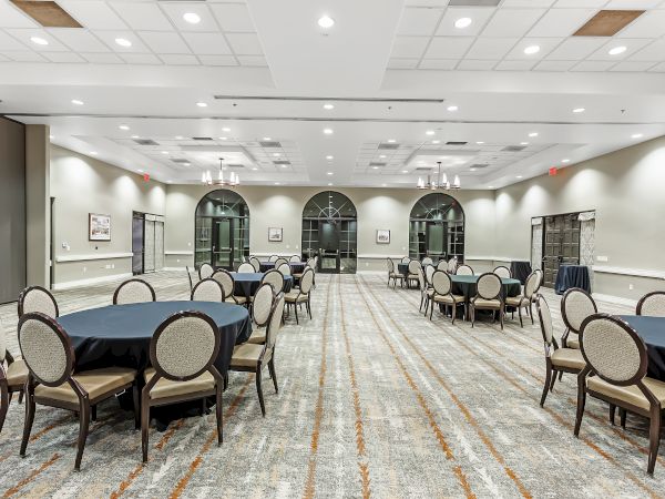 The image shows a spacious, well-lit conference or banquet room with round tables and chairs set up, and large windows along the back wall.