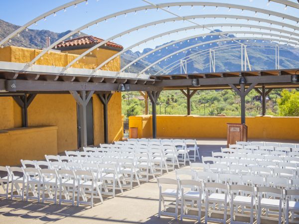 An outdoor setup with white chairs under a structure with curved beams, mountains in the background, and a wooden podium facing the seats.