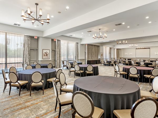 A spacious, well-lit event room with round tables and cushioned chairs, chandeliers, and large windows with drapes.