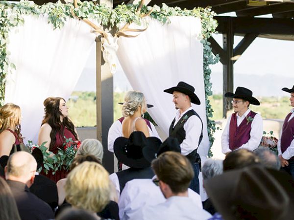 The image shows a wedding ceremony held outdoors. The bride and groom, along with bridesmaids and groomsmen, are dressed in semi-formal attire with hats.