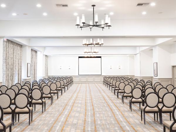 The image shows a conference room with rows of neatly arranged chairs, chandeliers, and a presentation screen at the front, ready for an event.