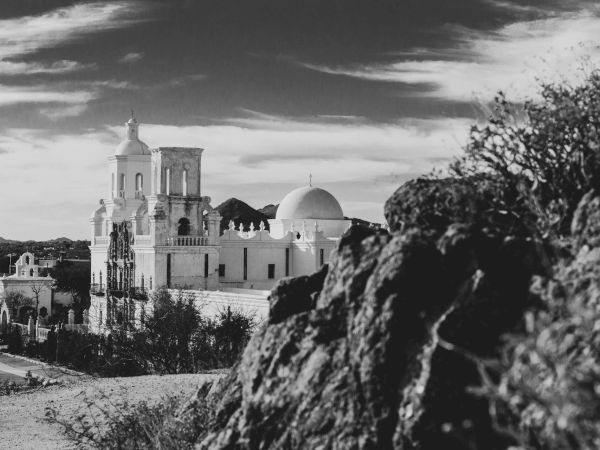 A black and white image featuring a historic mission-style building with domes and towers, set against a dramatic sky, partially obscured by rocky terrain.