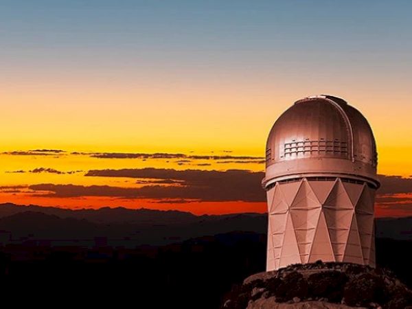 The image shows an observatory with a dome structure against a vibrant sunset sky with mountains in the background.
