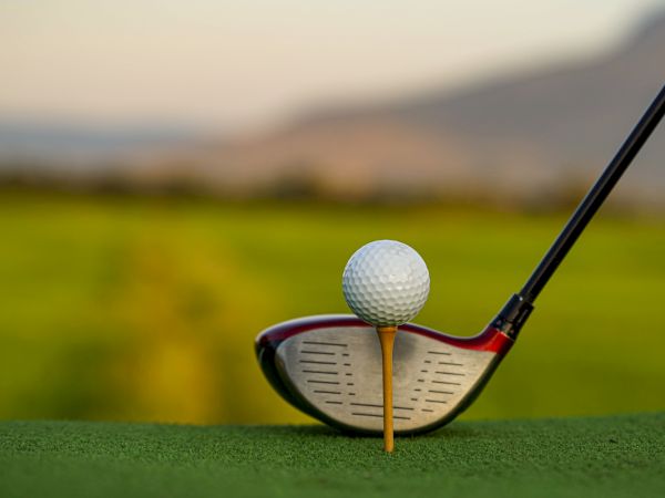 A golfer sets up for a shot, with a ball teed up, ready to be hit by the driver, against a grassy background and mountains in the distance.