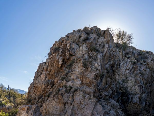 The image shows a large rocky formation with a bright blue sky overhead and some vegetation in the background, suggesting an arid environment.