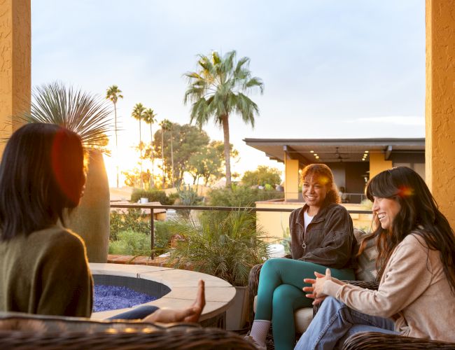 Three people chatting on a patio at sunset, with palm trees in the background.