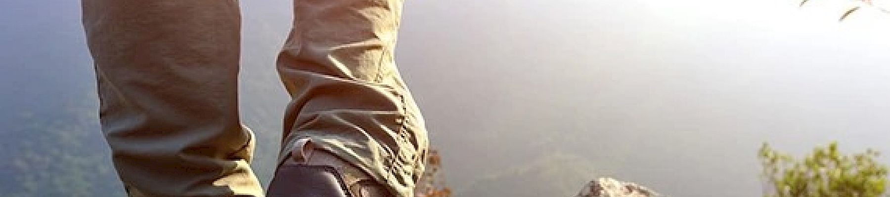 A person standing on a rocky cliff, wearing hiking boots and pants, with a mountainous landscape and bright sunlight in the background.