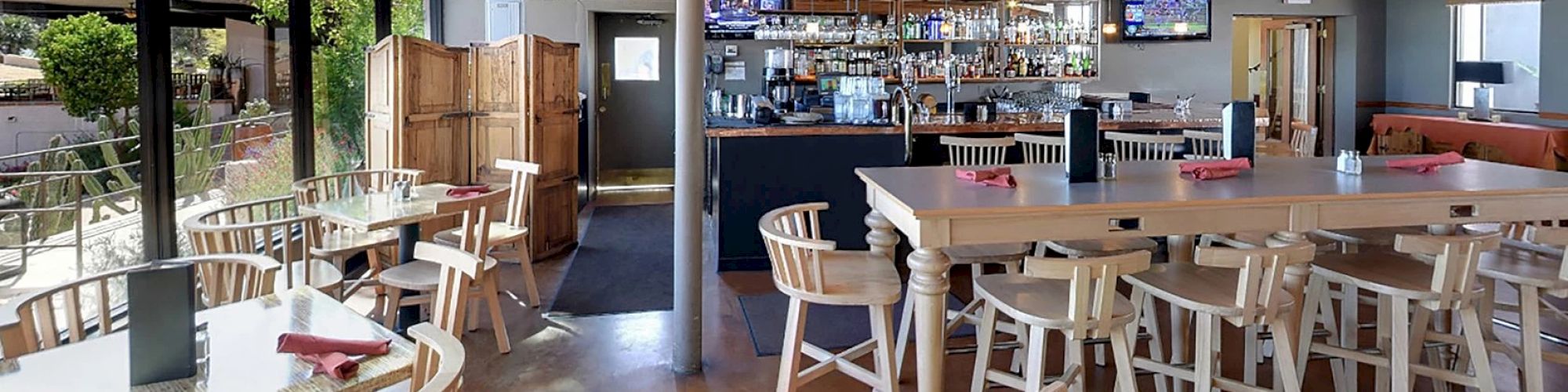 The image shows a bright, modern restaurant with wooden furniture, a bar area, and large windows providing an outdoor view.